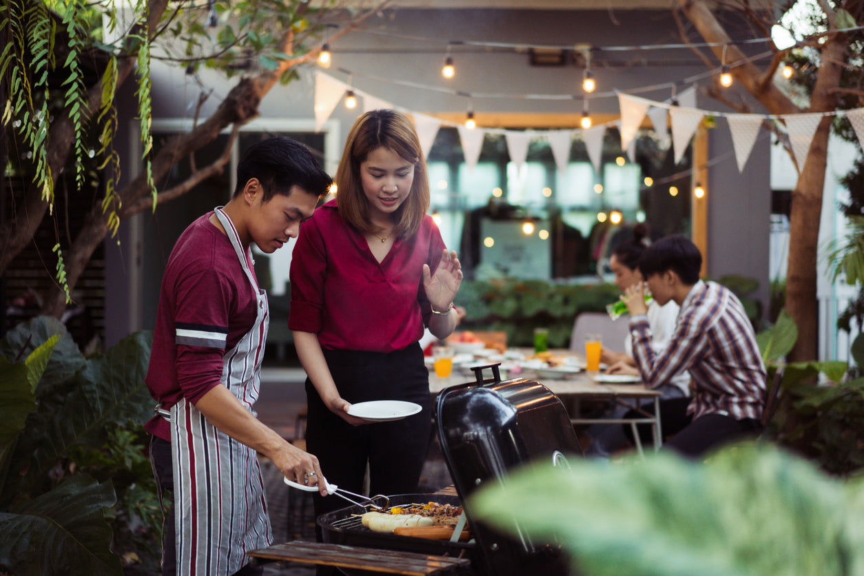 Barbecue etiquette for sensitive stomachs doesn’t have to be complicated! Image: A man and woman laughing over a barbecue as the man hands her a plate of meat. Behind them, several friends sit at a picnic table waiting to eat.